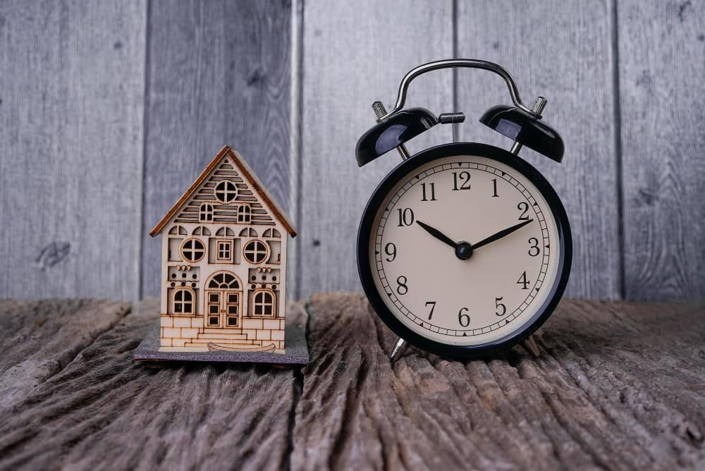 Un réveil analogique noir à clochettes est posé à côté d'une petite maquette de maison en bois sur une surface rustique, illustrant la discrétion des horloges espions