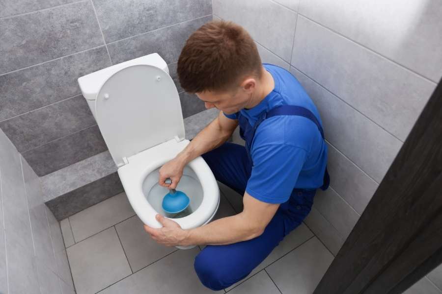 Plombier en combinaison bleue utilisant une ventouse pour déboucher ses WC dans des toilettes carrelées gris.