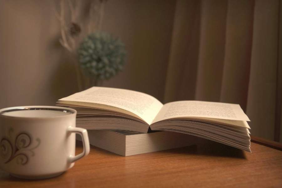 Gros plan chaleureux sur une table en bois avec un livre ouvert posé sur une pile et une tasse de café décorée, dans une ambiance feutrée.