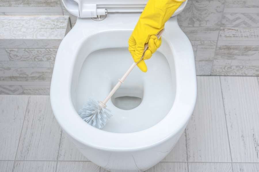 Une personne portant un gant de ménage jaune nettoie la cuvette en céramique blanche d'un WC à l'aide d'une brosse adaptée.